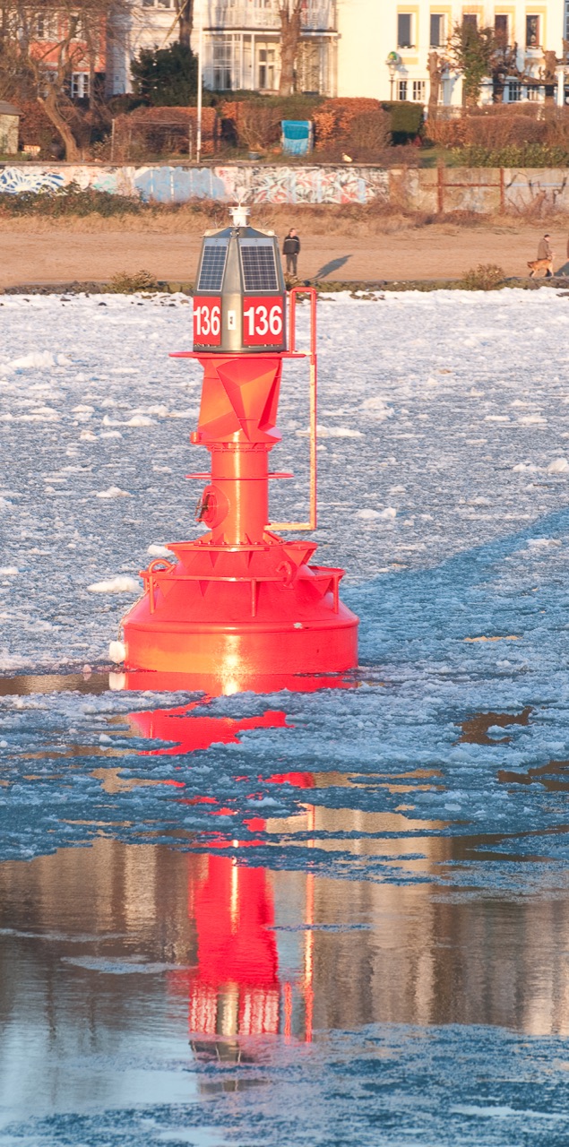 Red port-hand lateral buoy 136 among ice on the River Elbe, with a busy residential shoreline behind it.