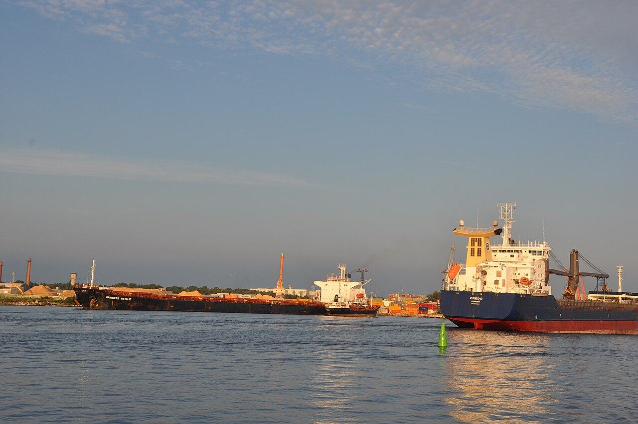 Small green IALA-A starboard-hand buoy in front of commercial ships and an industrial harbour at Riga.